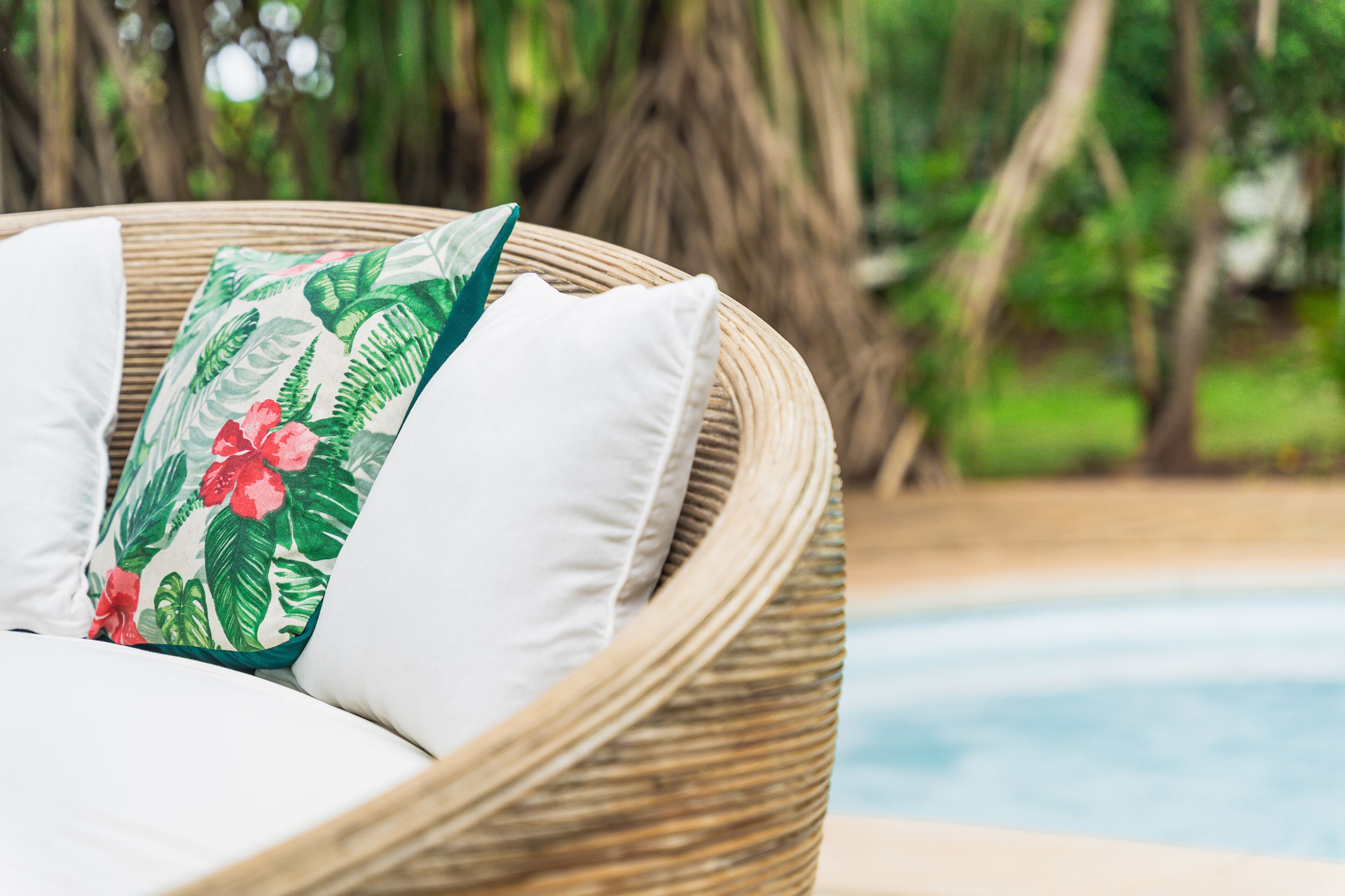 A wicker lounge chair with white cushions and a floral pillow, positioned near a pool with a blurred natural background.