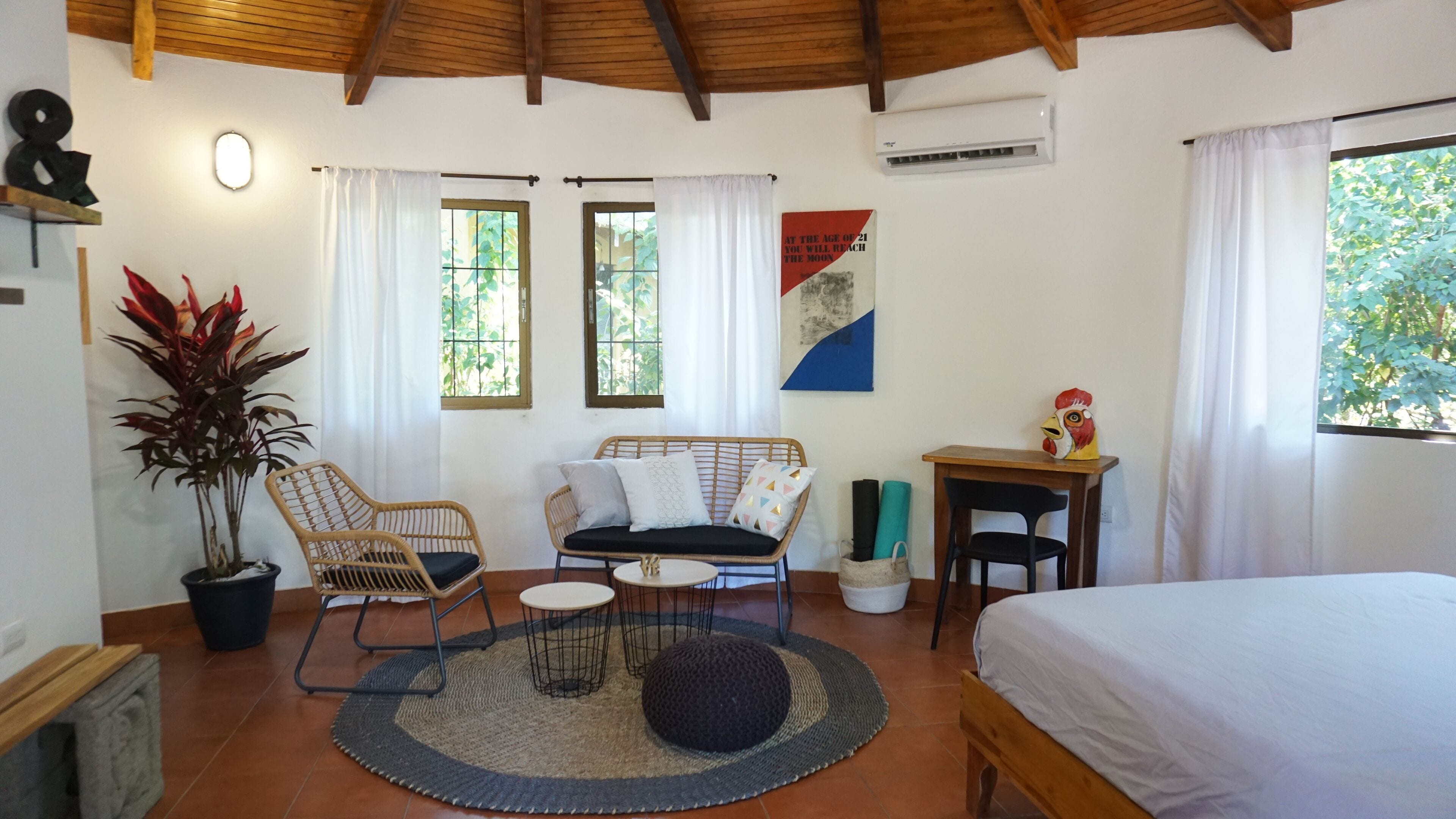 Ein gemütliches Zimmer mit Bett, einer Couch, einem kleinen Tisch und einem Sessel. Zwei Fenster mit Vorhängen, ein Blumentopf und eine Wand-Klimaanlage sind vorhanden.