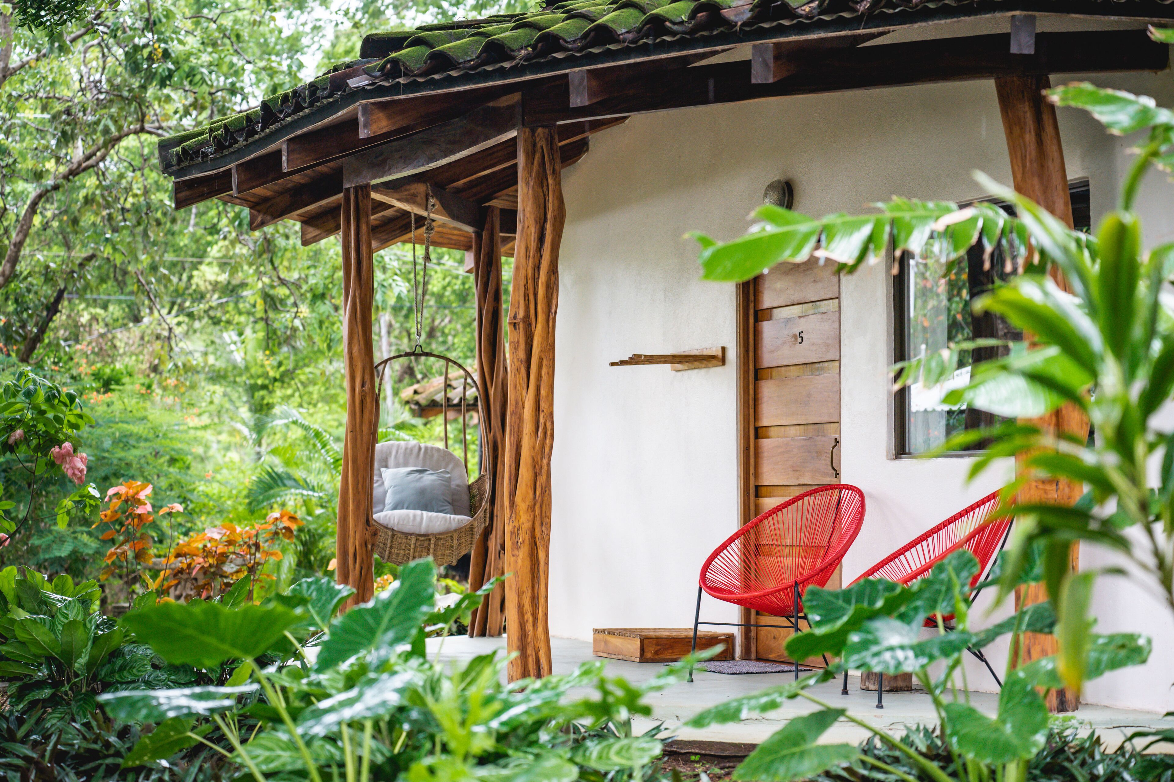 Une pergola en bois, avec un fauteuil suspendu et deux chaises rouges, se trouve devant un bâtiment blanc doté d’une porte en bois.
