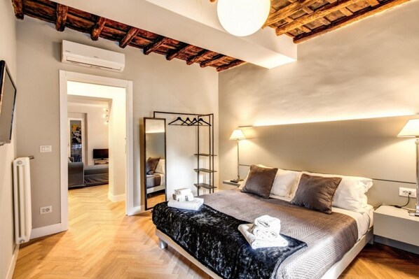 A modern bedroom with a wooden ceiling, a bed with a black and white duvet, bedside lamps, and a mirror with a clothing rack.