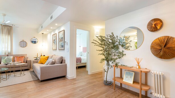 Een moderne woonkamer met een bank, salontafel en decoratieve spiegels. Er staat een plant in een glazen vaas en een klein houten bijzettafeltje met een fotolijstje.