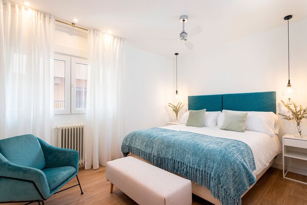 Un dormitorio moderno con cabecera color turquesa, una cama grande, un banco, una silla y una mesita de noche con lámpara.