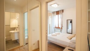 Ein Hotelzimmer mit Bett, Nachttisch, Spiegel, einem Fenster mit Vorhängen sowie einem Badezimmer mit Waschbecken und Spiegel.