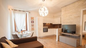 Ein modernes Wohnzimmer mit braunem Sofa, einem Esstisch mit Stühlen, einer Küchenzeile inklusive Fernseher und einer Wand mit Steinoptik.