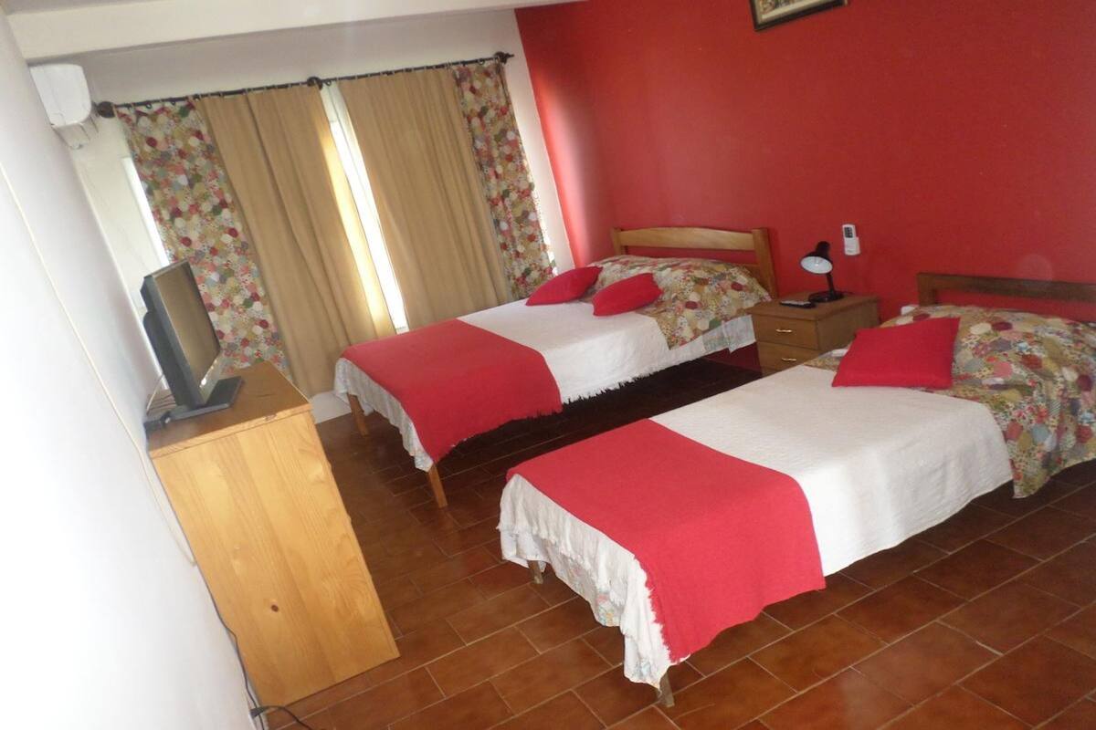 Una habitación con dos camas, cada una con un cojín rojo y ropa de cama blanca, una mesita de noche de madera con una lámpara y un televisor sobre un mueble.