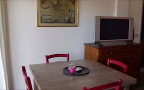 Un coin repas avec une table en bois, des chaises rouges, un téléviseur posé sur un meuble en bois et un tableau accroché au mur.