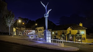 Escena callejera nocturna con una escultura destacada en el centro, un edificio con un letrero que dice "Earthy" y un cartel iluminado para "Paudy Old Irish Whiskey".
