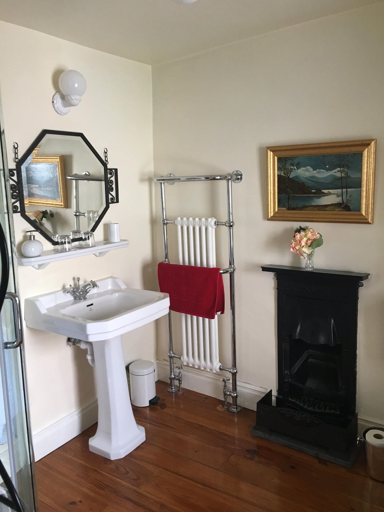 Une salle de bain avec un lavabo sur colonne blanc, un miroir avec un cadre décoratif, un radiateur, une cheminée noire et un tableau représentant un paysage, encadré.