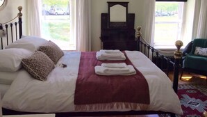 Una cama bien hecha con almohadas blancas y marrones, una manta roja y toallas dobladas. Se ve un aparador de madera con un espejo encima, y una silla verde en la esquina.