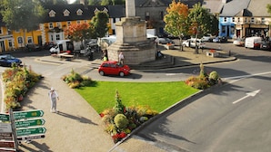 Una plaza de pueblo con un monumento, parterres de flores y señales direccionales.