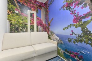 A room with a sofa, a mural of a coastal landscape, and a view of the sea.