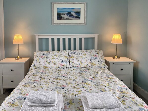 A bedroom with a bed, two bedside tables, two lamps, a framed picture on the wall, and two folded towels on the bed.