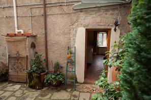 A rustic stone building with a doorway leading to a room with a tiled floor and a window. There are potted plants and a metal gate outside.