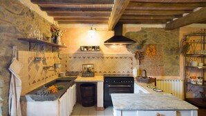 A rustic kitchen with a stone wall, wooden beams, a black stove, and a marble table. - Stone house with 4 double bedrooms and kitchen with fireplace (Boveglio)