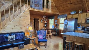 L’intérieur chaleureux d’un chalet comprend un escalier en bois, un salon avec un canapé et une table basse, un coin repas avec des chaises, une cuisine avec un réfrigérateur et un bar avec des tabourets.