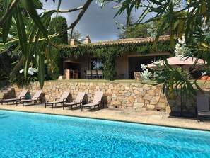 A poolside area with lounge chairs, a stone wall, and a building with a tiled roof.