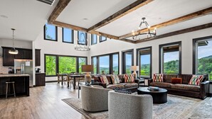 Un salón amplio con un gran ventanal con vistas a un paisaje exuberante, una zona de estar acogedora con chimenea, y una cocina moderna con armarios oscuros.