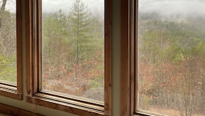 Una habitación con tres ventanas que dan a un paisaje boscoso con montañas y nubes.