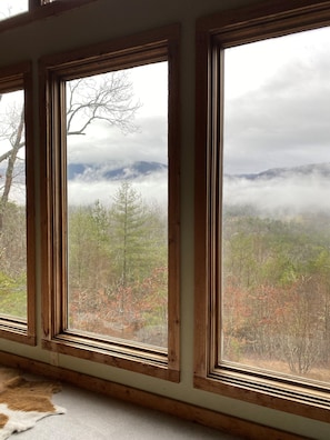Quarto com três janelas dando vista para uma paisagem arborizada com montanhas e nuvens.