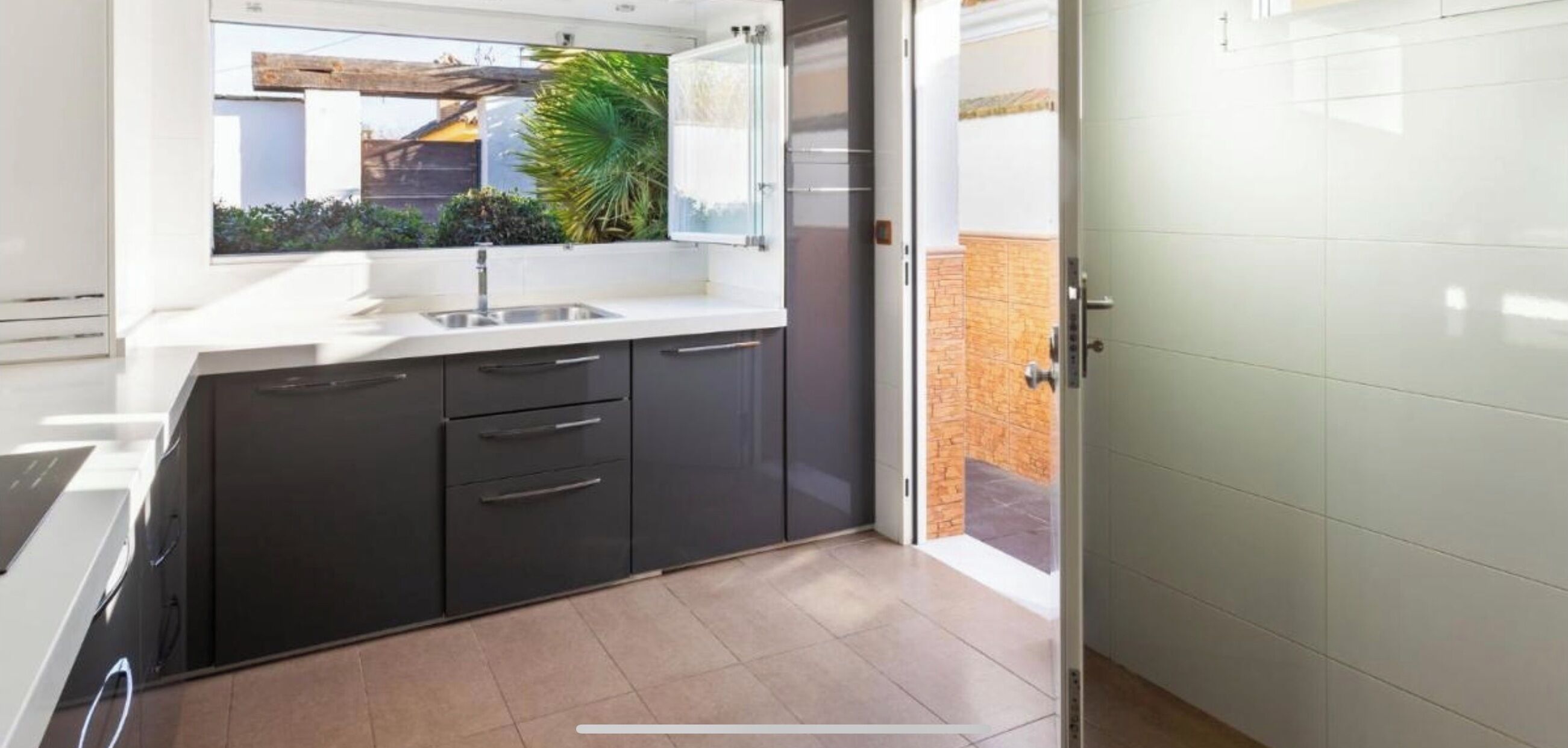Una cocina moderna con armarios oscuros, encimera blanca y ventana con vistas a un jardín.