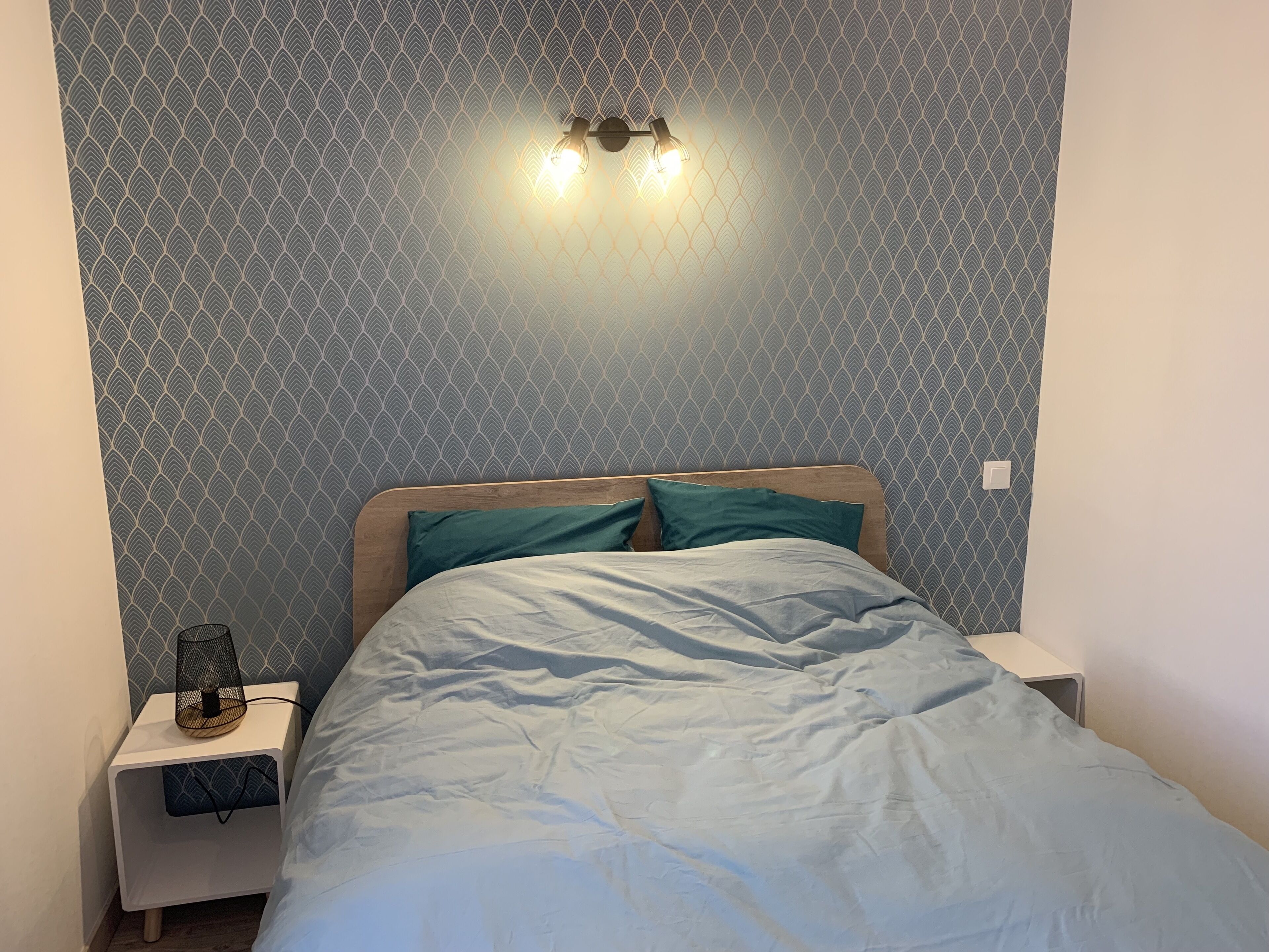 Un dormitorio con papel tapiz estampado, una cama con ropa de cama color turquesa y una mesita de noche con una lámpara.