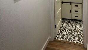A hallway with a white door leading to a bathroom. The bathroom has a white sink, a mirror, and a patterned floor. There are four black-framed mirrors on the wall.