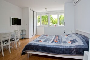 Quarto com cama, escrivaninha, cadeira e televisão. Há uma janela com vista para árvores.