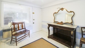 Una habitación con un banco de madera, una consola de madera oscura con un espejo de marco dorado y una puerta blanca con un ventanal con persianas.