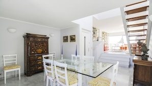Un área de comedor con una mesa de vidrio y sillas blancas, un armario de madera, y una escalera que lleva a un balcón.
