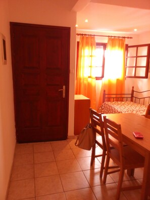 Une pièce avec une porte en bois, un lit recouvert d’une couvre-lit à motifs, une table avec des chaises et une fenêtre avec des rideaux.