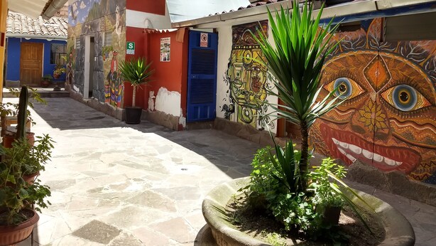 A courtyard with a tiled floor, potted plants, and colorful murals on the walls. - Chelitos Backpacker (Cusco)