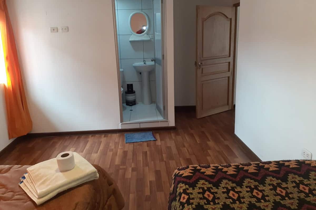 A hotel room with a wooden floor, a bed with a patterned cover, a small table with a towel and a bottle, a bathroom with a sink and mirror, and a wooden door.