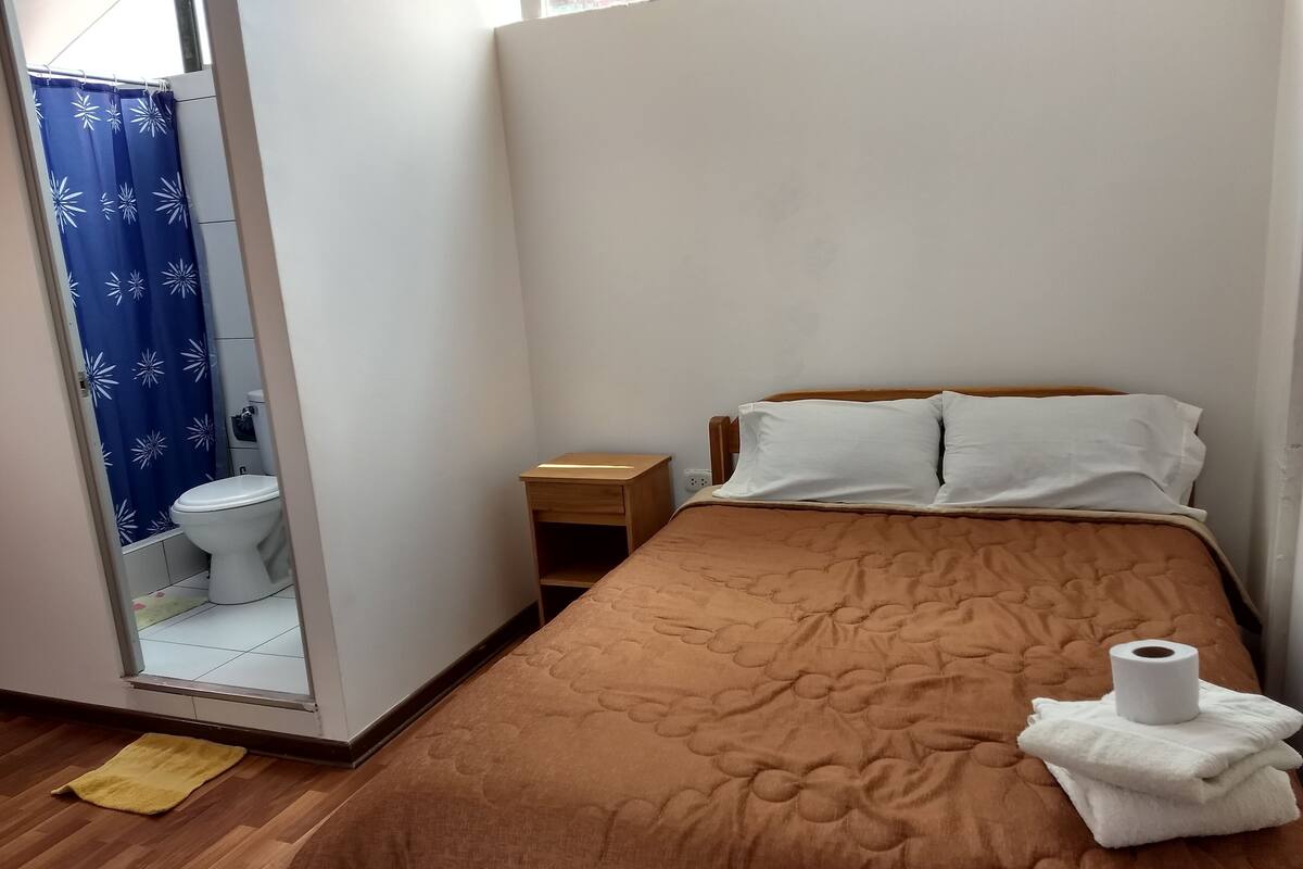 A bedroom with a bed, a wooden nightstand, a white pillow, and a roll of toilet paper on the bed.
