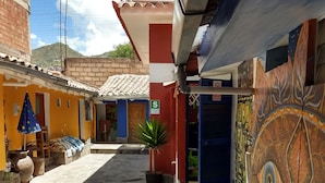A narrow alleyway with a tiled floor, a small plant, and a colorful mural on the right wall. - Chelitos Backpacker (Cusco)