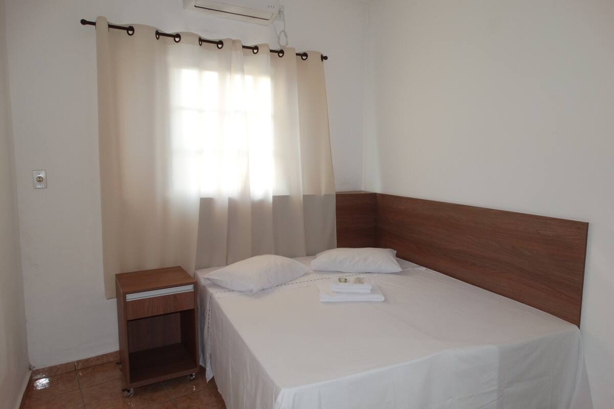 A neatly made bed with white linens, a wooden nightstand, and a window with sheer curtains.