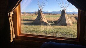 Une vue par la fenêtre sur deux tipis installés dans un champ herbeux, avec des montagnes en arrière-plan.