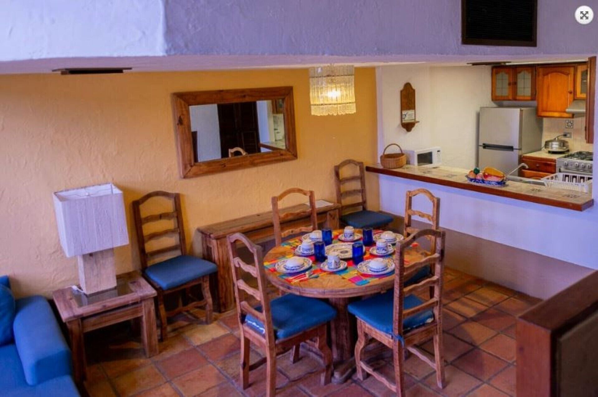 A dining area with a round table set for four, surrounded by wooden chairs with blue cushions. A small wooden side table with a lamp is to the left. The room has a tiled floor and a kitchen area with a refrigerator and stove in the background.