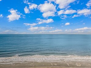 Une plage à l’eau d’un bleu limpide et au rivage sablonneux, sous un ciel d’un bleu éclatant parsemé de quelques nuages.