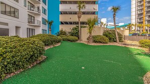A courtyard with artificial green turf, surrounded by dense hedges and palm trees, with modern buildings in the background. - The Palace 105 (Myrtle Beach)