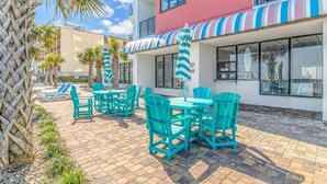 A patio area with turquoise chairs and tables, a striped awning, and a building with balconies and large windows. - The Palace 105 (Myrtle Beach)