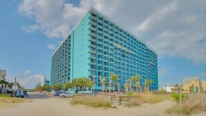Un hotel alto de color azul, con varios pisos y balcones, situado cerca de una playa con palmeras y un camino de arena.