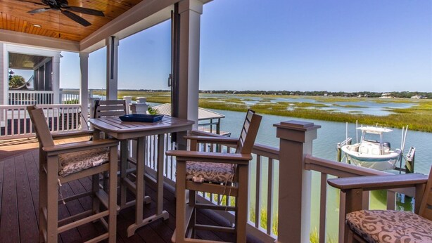 A covered outdoor area with a table and chairs, a boat docked nearby, and a view of a water body and distant landscape. - Channel Marker (Murrells Inlet)