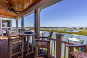 A covered outdoor area with a table and chairs, a boat docked nearby, and a view of a water body and distant landscape. - Channel Marker (Murrells Inlet)