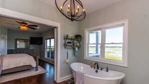 A spacious bathroom with a freestanding bathtub, a ceiling fan, and a large window offering a view of the outdoors. - Channel Marker (Murrells Inlet)