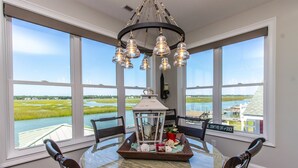 A dining area with a round glass table, four chairs, a chandelier, a lantern, and a view of a water body through large windows. - Channel Marker (Murrells Inlet)