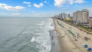 Une plage avec une longue étendue de sable, une jetée visible au loin et plusieurs immeubles de grande hauteur le long de la côte.