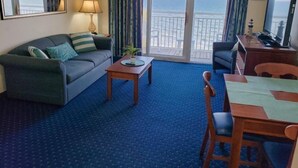 A hotel room with a sofa, a small table, a desk with a chair, a TV, and a view of the ocean through the windows. - Bay Watch 1438 (North Myrtle Beach)