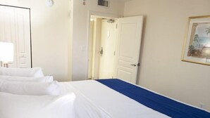 A hotel room with a large bed, white linens, a blue accent on the bed, a door, and a framed picture on the wall. - Bay Watch 1438 (North Myrtle Beach)