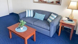 A room with a blue patterned carpet, a grey sofa with striped pillows, a wooden coffee table with a plant, a bedside table with a lamp, and a mirror on the wall. - Bay Watch 1438 (North Myrtle Beach)