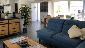 Een moderne woonkamer met een blauwe bank, houten kasten, een koffieapparaat en een eethoek met gele stoelen.